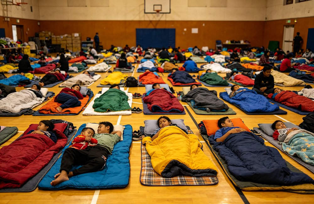 Wide angle view of emergency evacuation center in gymnasium with families resting on colorful horizontally laid sleeping bags, natural daylight from windows, disaster relief shelter, Asian people