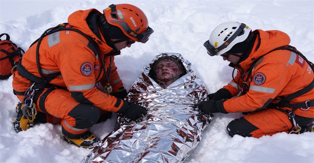 Wide view of avalanche survivor wrapped in warm blanket by rescuers showing post-rescue thermal care