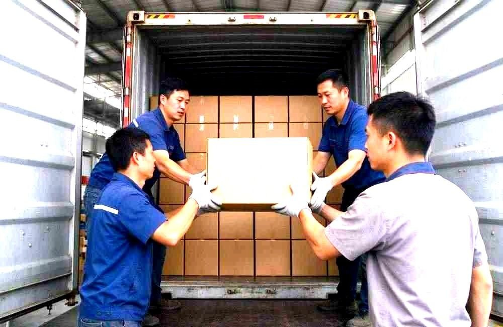 Wide view of Asian warehouse workers loading cartons of bulk blankets into a shipping container at a loading dock, organised pallets, daylight