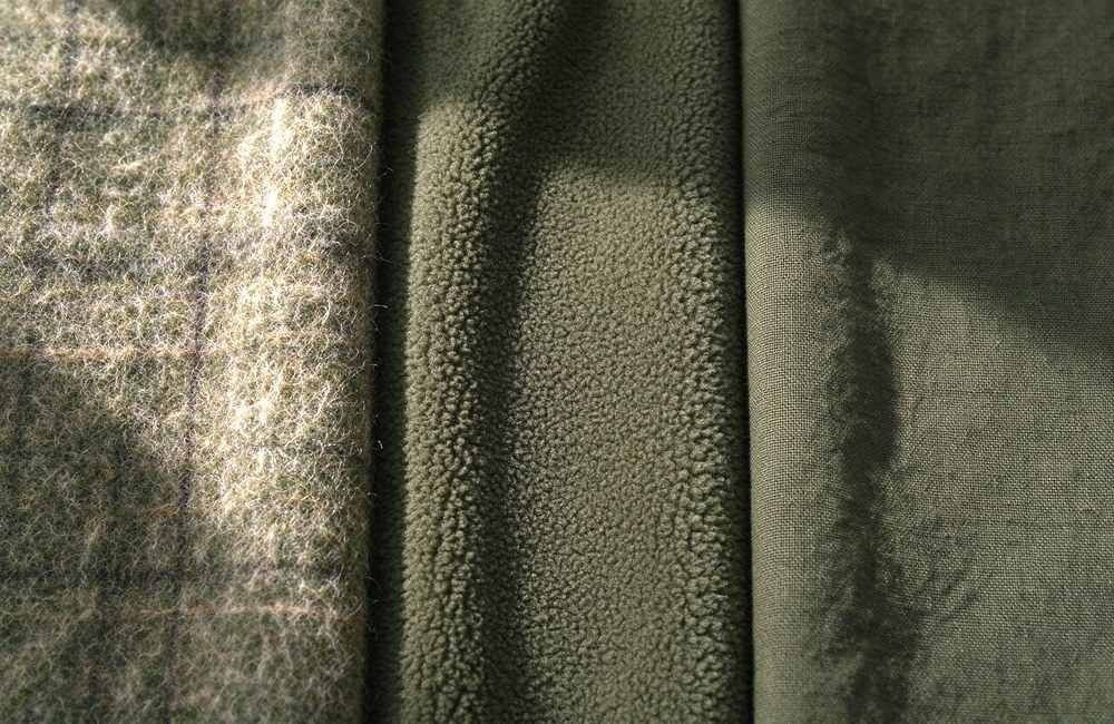 Close-up shot side by side comparison of olive green wool blend, fleece and cotton blanket fabrics showing different texture details